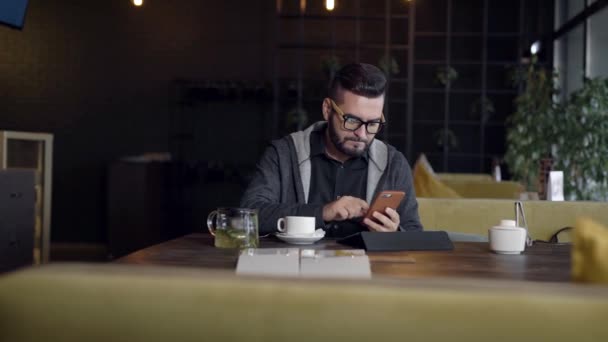 Hombre hipster guapo está escribiendo en la pantalla del teléfono inteligente sentado en el restaurante durante el día, tableta en frente — Vídeo de stock