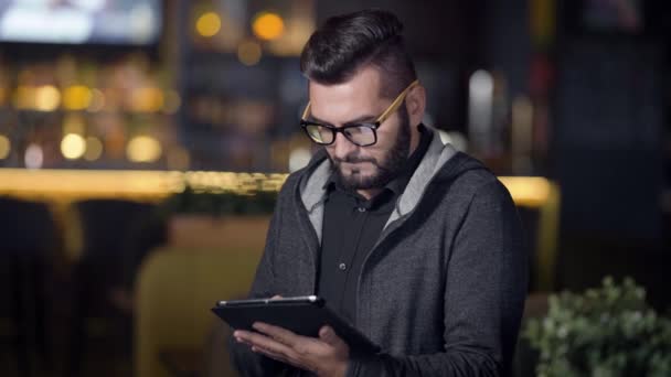 Hombre adulto con peinado de moda y gafas en la cara está navegando por Internet por tableta sentado en el bar por la noche — Vídeo de stock