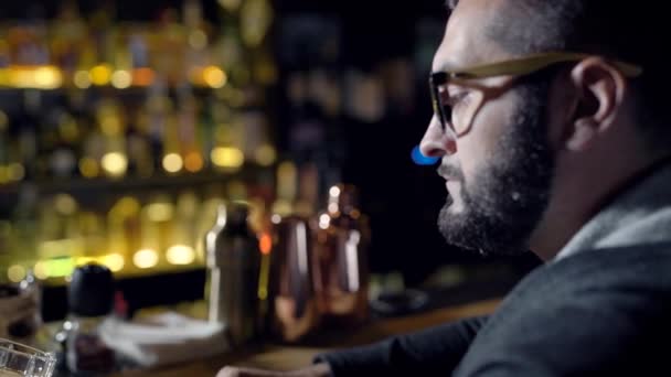 Solo hombre adulto está bebiendo cerveza sentado en un bar por la noche, degustación de cócteles, vidrio giratorio en un mostrador — Vídeos de Stock