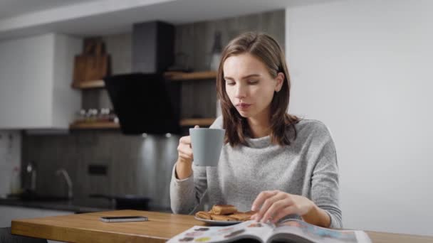 young woman is reading magazine and drinking coffee in morning in holiday, sitting on her kitchen in apartment