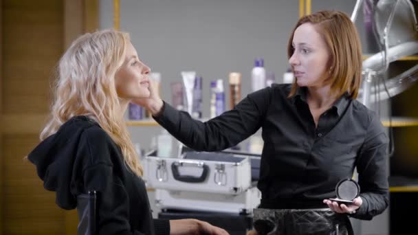 Vrouwelijke make-up artiest bedekt gezicht van jonge blonde vrouw door cosmetische poeder, penseel, met schoonheidssalon — Stockvideo