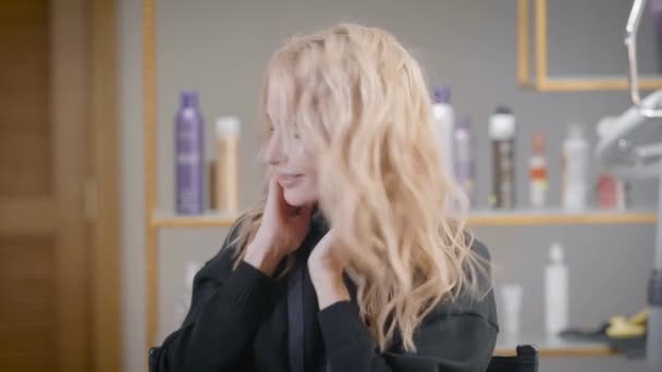 Молодая красивая блондинка демонстрирует свою прическу в парикмахерской, машет головой и замками — стоковое видео