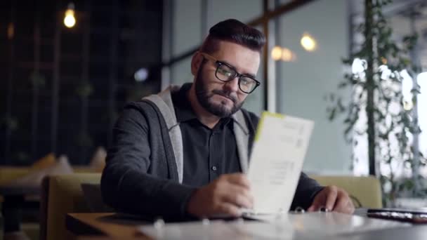 Elegante chico con barba en gafas llegó en el restaurante y la elección de la comida en el menú . — Vídeos de Stock