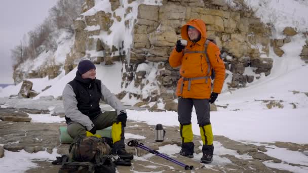 Dois caminhantes pararam na margem do rio para descansar e beber um chá quente durante a caminhada de inverno . — Vídeo de Stock