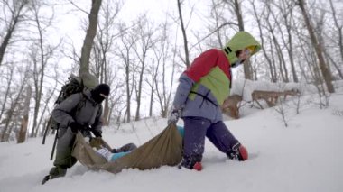Kışın ormanda yaralı bir kadını sedyede sürükleyen trekking ekipmanı olan iki adamın yan görünümü.