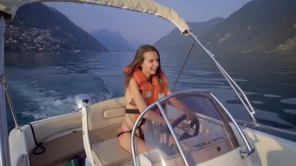 Hermosa morena en un chaleco salvavidas conduce un pequeño barco a motor. La chica sonríe, le gusta la vista alrededor, el lago y las montañas. Vacaciones verano — Vídeos de Stock