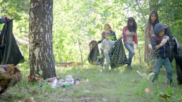 Skupina žen a dětí sbírá odpadky v lese. Dobrovolníci sbírají plastové a jiné odpady v černých plastových sáčcích. Za životní prostředí jsou odpovědni lidé.