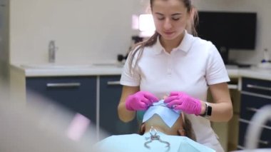 Diş hekimi maske eldiveni ve mavi üniforma giyiyor. Hasta dişlerini diş araştırması ve arka planda tıbbi ekipmanlarla dolu bir kutuda ayna ile inceliyor.