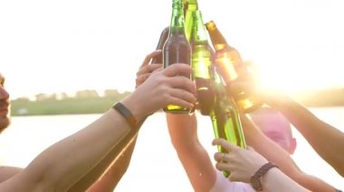 Bira şişeleriyle kadeh kaldıran ve gün batımında müzik eşliğinde dans eden bir grup etnik kökenli genç, gün batımında bira içen arkadaşlar.