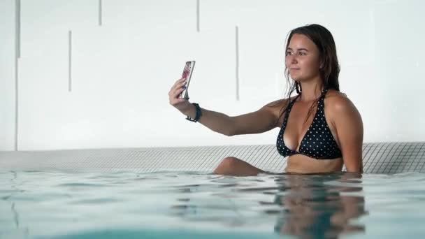 リゾートのスパでスマホで自撮りしてる美少女 携帯電話を手に夏には水の中でリラックスして 水着でブルネット — ストック動画