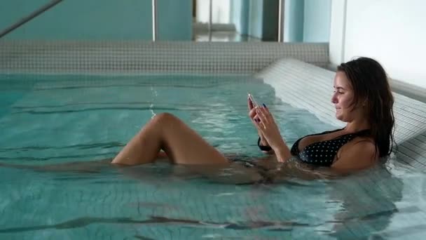 かなりブルネットはプールでスマートフォンを使用しています 水着姿の女の子が休んでいる — ストック動画