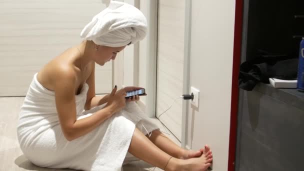 머리에 수건을 샤워를 마치고 아름답고 여성은 스마트폰을 사용하고 스크롤 페이지를 — 비디오