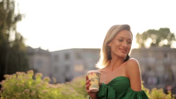 一个金发碧眼的女孩在镜头前笑得紧紧的 有树木的城市景观 手里拿着纸杯咖啡 — 图库视频影像