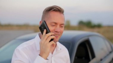 Yakışıklı gülümseyen iş adamı arabanın yanında duruyor ve cep telefonu kullanıyor..