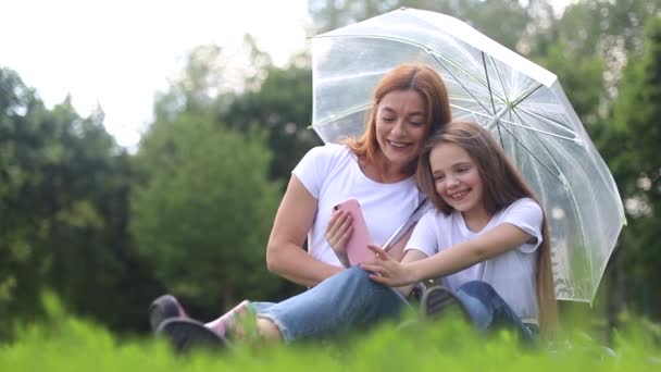 Feliz madre sonríe y juega con su hija en el parque escondiéndose de la lluvia — Vídeos de Stock