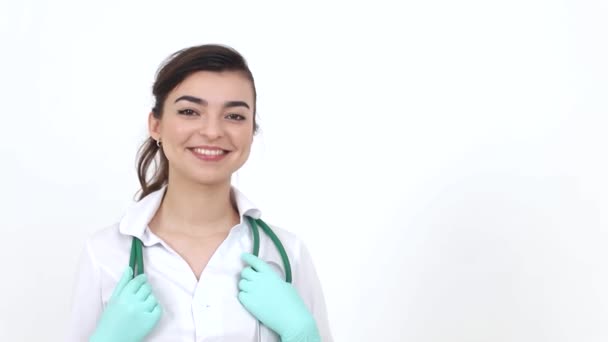 Eine junge Ärztin in weißem Arztkittel mit Stethoskop blickt in die Kamera. Lächelnde Ärztin, mittlere Einstellung auf weißem Hintergrund — Stockvideo