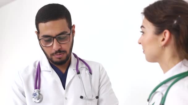 Dois jovens médicos, um homem e uma mulher de casacos brancos, conversam usando um tablet digital . — Vídeo de Stock