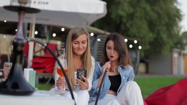 二人の女の子はフックを吸うと写真を見てください 若い人たちは日没に座っている 湖のそばに座っている — ストック動画
