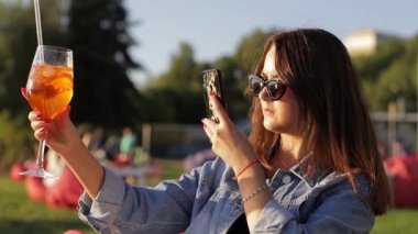 Bir kadın portakal kokteyli içer ve akıllı telefonuyla fotoğrafını çeker.