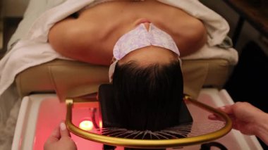 Göz maskesiyle spa yatağında yatan bir kadın, parlak sağlık ışığı altında kafa derisini yüzüyor..