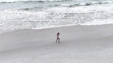 Okyanusun yakınındaki kumlu bir plajda tek başına koşan atletik bir kadın. Drone videosu.