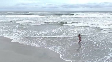 Mayo giymiş genç bir kadının kumlu sahil boyunca sığ suda yürüdüğünü gösteren hava aracı videosu..