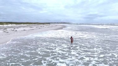 Bikinili bir kadının kumsalda yürüyüp dalgaların keyfini çıkardığı drone görüntüleri. Manzaralı yaşam tarzı ve özgürlük teması.