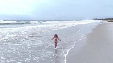 Mayo giymiş genç bir kadının kumlu kıyı şeridinde sığ suda yürüdüğünü gösteren drone videosu. Rahatlatıcı yaşam tarzı ve yaz tatili konsepti.