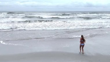 Bulutlu gökyüzünün altında okyanus dalgalarıyla kumlu sahilde gezinen bir kadının insansız hava aracı görüntüsü. Seyahat ve doğa projeleri için rahatlatıcı yaşam tarzı sahnesi.