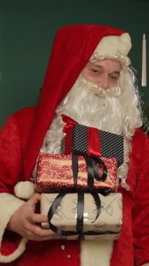 Noel Baba, güzel dekore edilmiş bir ağacın önünde, neşeli ve neşeli bir Noel atmosferi yaratarak, mutlu bir şekilde paketlenmiş bir Noel hediyesi sunar. Dikey video.