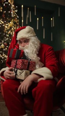 Noel Baba kostümü giymiş bir adam süslü bir Noel ağacının yanında elinde neşeyle renkli hediyeler tutuyor ve bayram sezonunun şenlik ruhunu somutlaştırıyor. Dikey video.