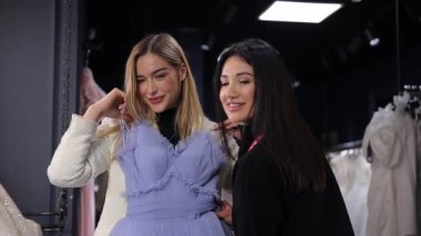 Moda mağazasında şık bir elbise provası sırasında gülümseyen ve kameraya poz veren iki kadın.