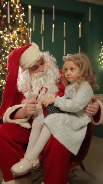 Noel Baba 'nın kucağında oturan, elinde şeker bir değnekle parıldayan gözlerle gülümseyen ve süslü bir Noel ağacının yanında mutlu bir şekilde sohbet eden tatlı küçük kız. Dikey video.