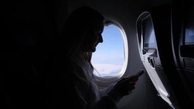 Uçak penceresinin yanında oturan bir adamın silueti, uçuş sırasında dışarıdaki bulutlara bakarken bir telefon tutuyor. Seyahat, teknoloji ve havacılık kavramı.