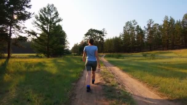 Chica en pantalones cortos y camiseta azul caminando por un camino rural al atardecer — Vídeos de Stock