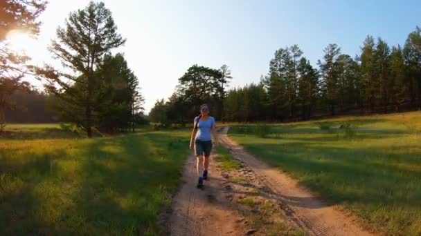 Chica en pantalones cortos y gafas azules caminando por un camino rural al atardecer — Vídeos de Stock