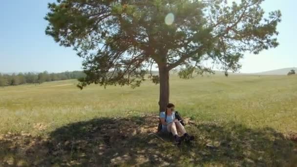 Chica sentada bajo un árbol, descansando a la sombra, dibujando algo en el álbum — Vídeos de Stock