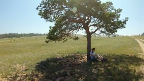 Mädchen sitzt unter einem Baum, ruht im Schatten und schreibt etwas in ein Notizbuch — Stockvideo