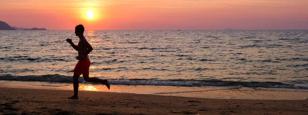 Sylwetka młodego mężczyzny biegającego po plaży o wschodzie słońca ze słońcem w tle. Zachowanie sprawności i zdrowy sztandar — Zdjęcie stockowe