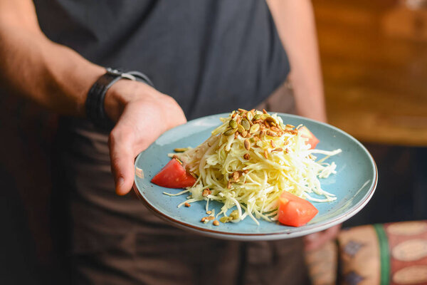 Cabbage salad with sunflower seeds and tomatoes. Served in a blue plate. In hand of a waiter. Restaurant service.