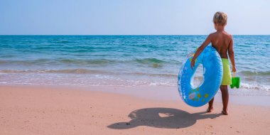 Mavi ve sarı bir havuz elinde yüzerek denizin kenarında duran mutlu küçük çocuk. Boşluk afişini kopyala.