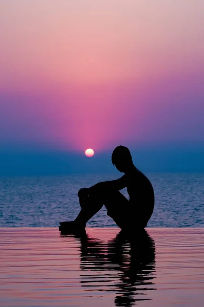 Мальчик сидит в бассейне и наслаждается закатом. Человеческий силуэт над закатом солнца. Бассейн по вечерам . — стоковое фото