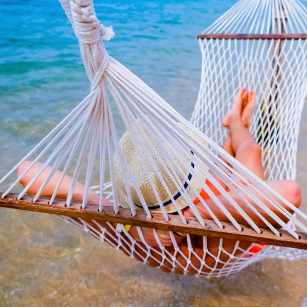 Boy with sandy feet lying in a hammock at the beach. Cozy hammock on the tropical beach by the sea. Peaceful seascape. Beautiful blue sky over calm sea with sunlight reflection. Copy space banner.
