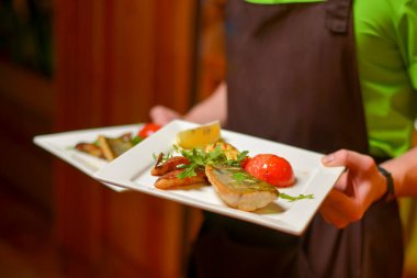 Balık tabağı - sebzeli kızarmış balık filetosu beyaz tabakta servis ediliyor. Garson akşam yemeğiyle birlikte bir tabak tutuyor. Restoran servisi, restoranda yemek yemek. Garson işte..