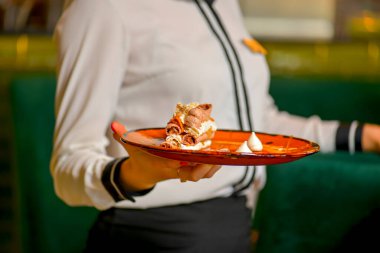 Waiter in white uniform, shirt, bringing the ordered dessert and in a cafe or restaurant. Piece of layered vanilla honey cake on a plate. Restaurant service. clipart