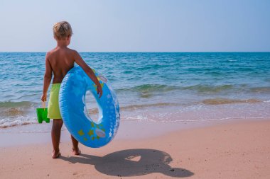 Mavi ve sarı bir havuz elinde yüzerek denizin önünde duran mutlu küçük çocuk. Yaz tatili, seyahat, turizm ve tatil konsepti. Boşluk afişini kopyala.