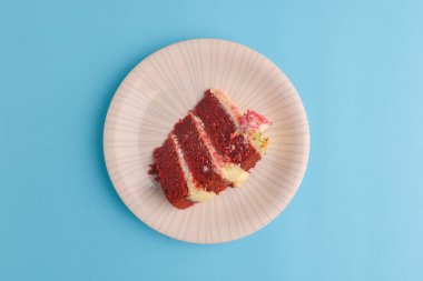 Bir dilim beyaz kremalı kırmızı kadife pasta ve üstünde küçük bir gül çiçeği. Parlak pastel mavi arka planda beyaz bir tabakta servis edilmiş. Lezzetli tatlı. Geleneksel kırmızı kadife kek tarifi.