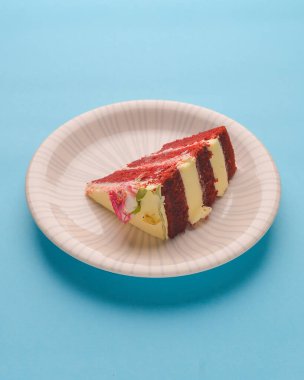 Bir dilim beyaz kremalı kırmızı kadife pasta ve üstünde küçük bir gül çiçeği. Parlak pastel mavi arka planda beyaz bir tabakta servis edilmiş. Lezzetli tatlı. Geleneksel kırmızı kadife kek tarifi.