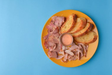 Dilimlenmiş jambon eti ve pastel mavi arka planda sarı bir tabakta servis edilen sosla servis edilen ekmek dilimleri..