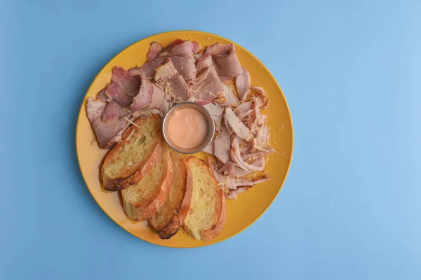 Sliced ham meat and cut pieces of bread served with sauce on a yellow plate over pastel blue background. Delicious snack, light healthy food, sandwich.